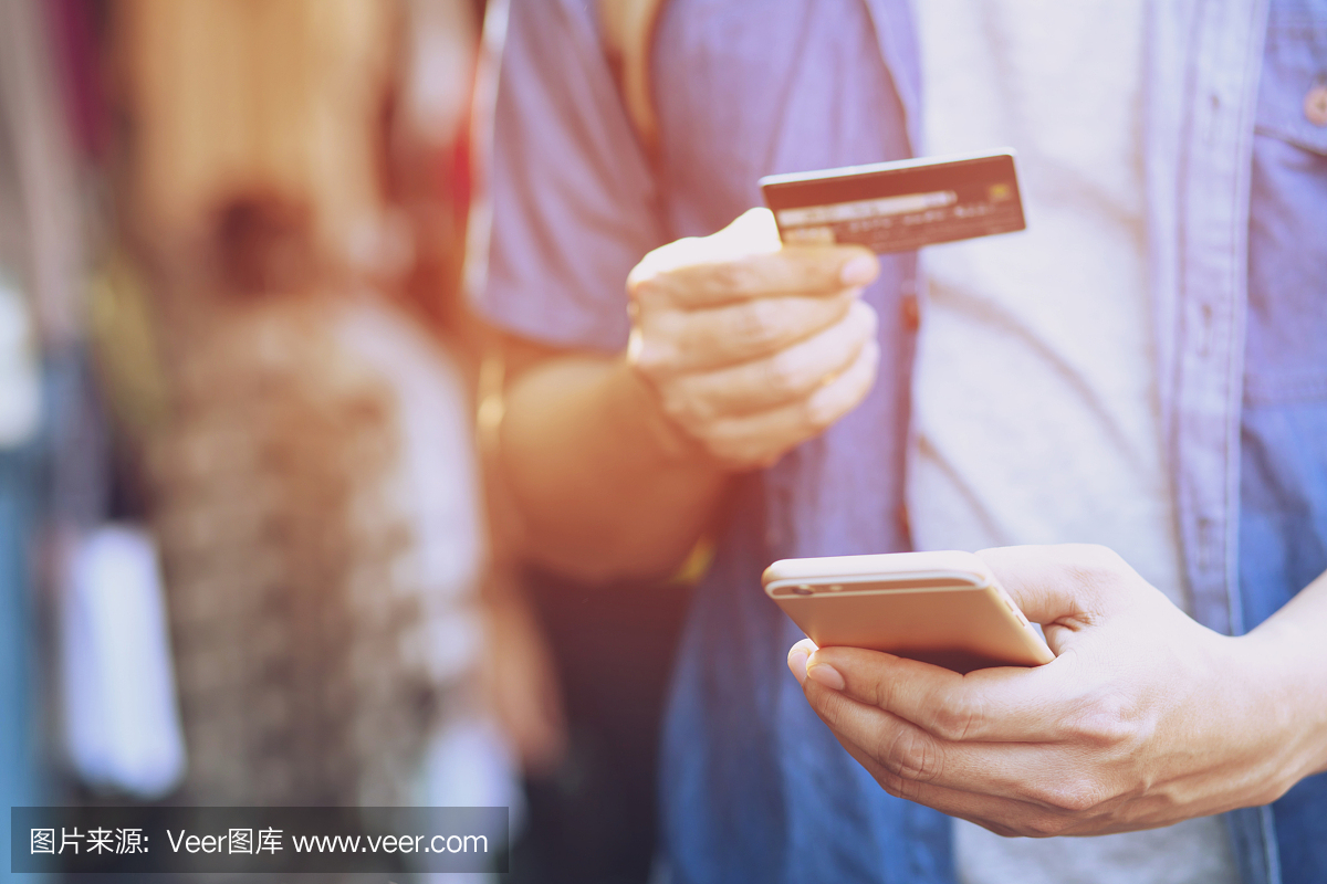 一名年轻男子手持信用卡,使用手机站在服装店门前。网上购物购买销售或支付。